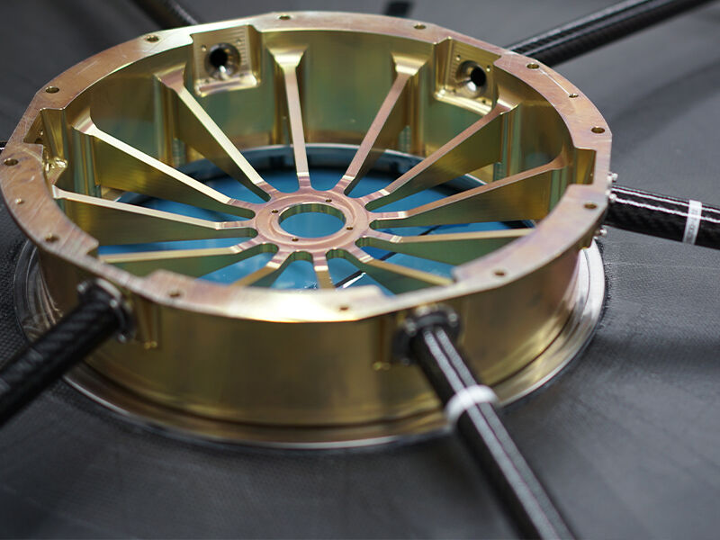 Image shows the underside of the deformable reflector. It is a closeup image of the center insert, a metallic component with spokes. There are small carbon fiber tubes extending outward from the exterior surface of the component. The carbon fiber surface of the back of the reflector is visible.